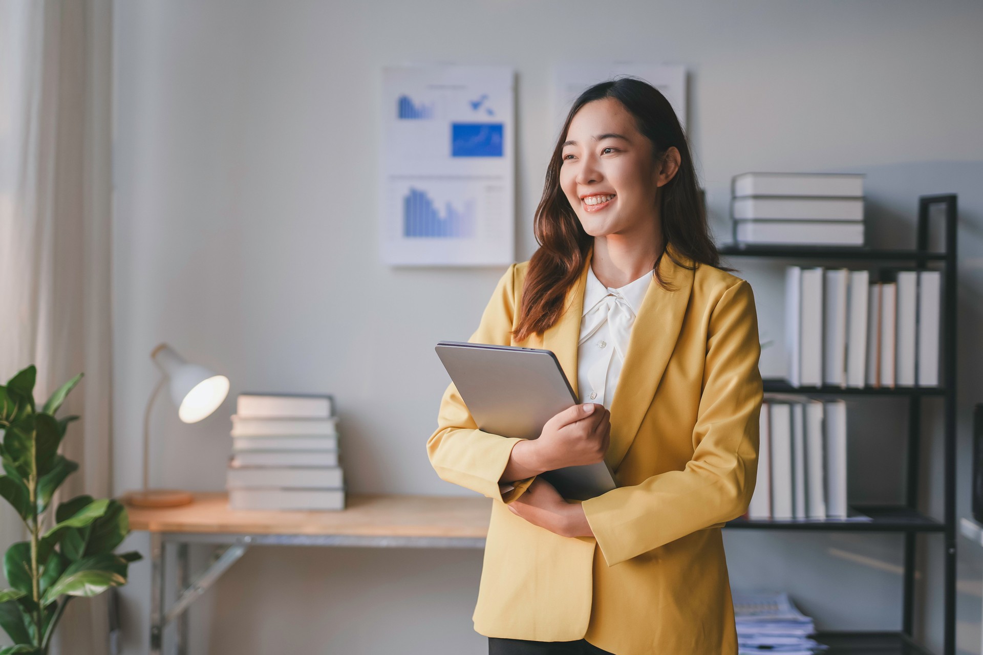 Young businesswoman is holding a laptop while looking out the window and smiling in her bright office