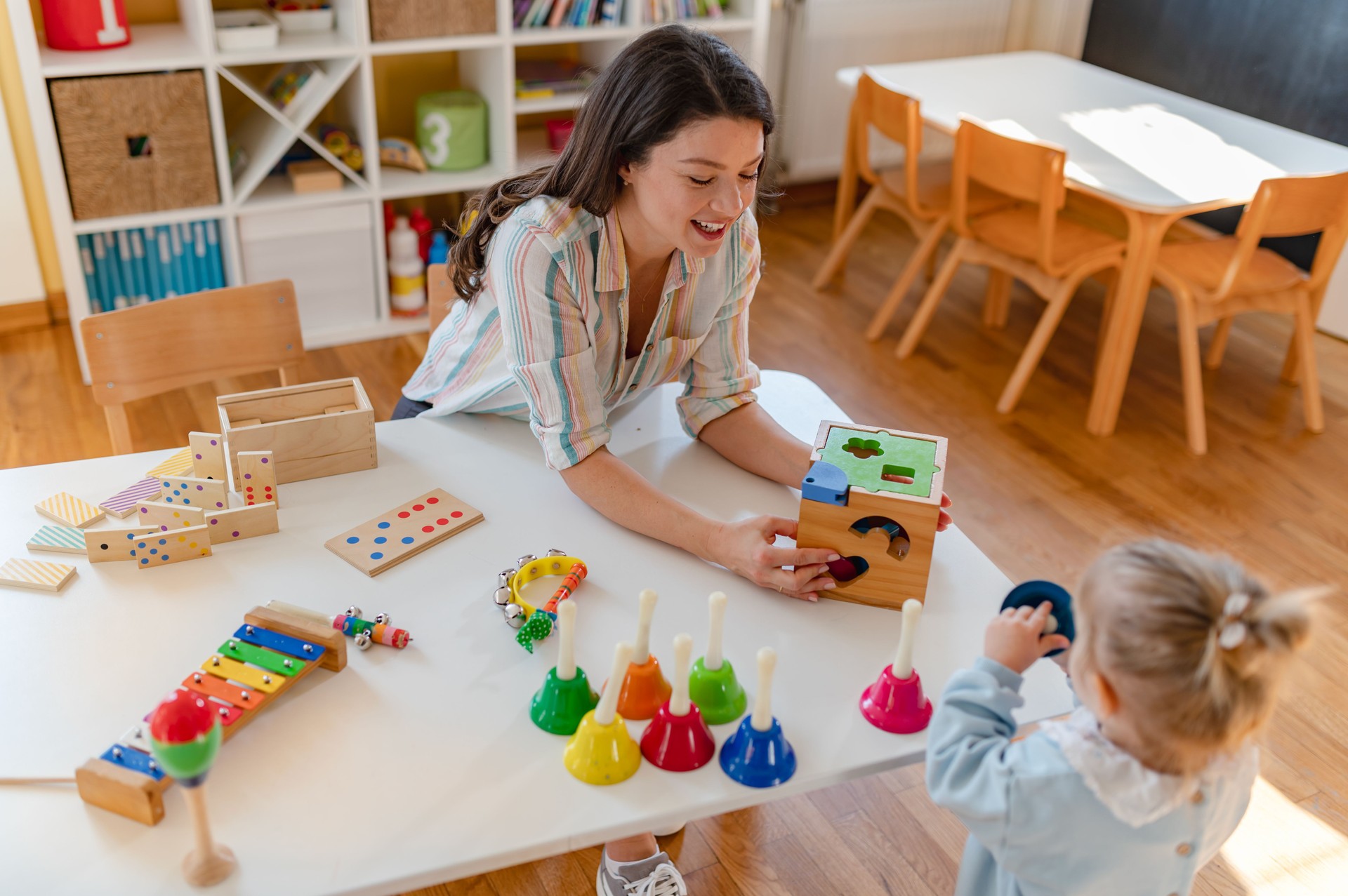 A young kindergarten teacher and a little girl happily play with wooden educational toys in a bright classroom, engaging in early learning activities that promote cognitive and motor skill development.